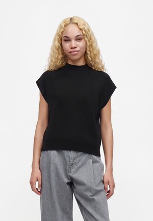 Jeune femme aux longs cheveux blonds bouclés portant un haut noir sans manches et un pantalon gris ample, debout devant un fond blanc uni.