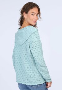 Himmelblaues, gestricktes Hoodie mit einem Spitzenmuster, gewelltem Saum und langen Ärmeln, getragen von einer Person, die sich leicht nach hinten dreht.