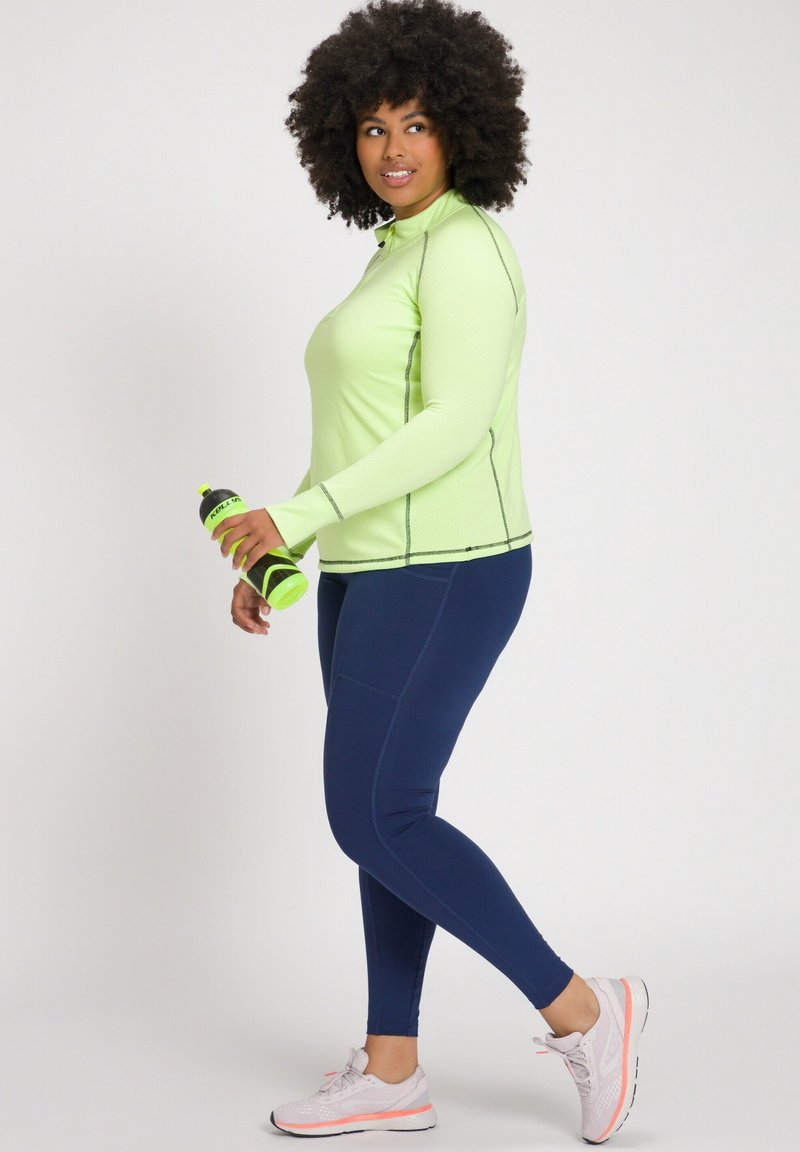 Langärmliges, hellgrünes Sportoberteil mit einem strukturierten Muster, kombiniert mit marineblauen Leggings. Das Model hält eine grüne Wasserflasche und trägt Schuhe mit pinken Akzenten.