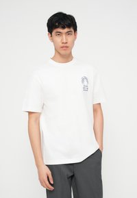 Νέος με μαύρα μαλλιά, που φοράει λευκό μπλουζάκι με μικρό λεπτομερές σχέδιο στο στήθος και γκρι παντελόνι, που στέκεται μπροστά σε απλό φόντο.