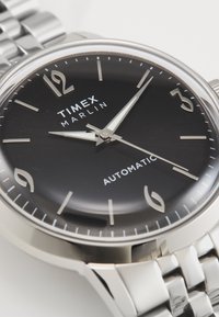 Silberne Automatikuhr mit schwarzem Zifferblatt, silberfarbenen Stundenmarkierungen und einem Armband aus Edelstahl. Verfügt über eine runde Form und eine polierte Oberfläche.