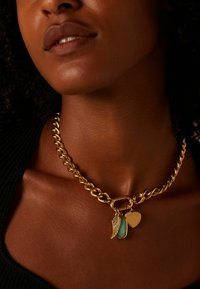 Collier en chaîne dorée mettant en avant un design de maillons entrelacés, agrémenté de trois pendentifs : une aile, un cœur et un pendentif de couleur turquoise.