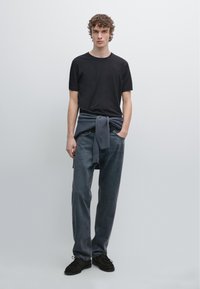 Sort t-shirt med korte ærmer kombineret med mørkegrå jeans. En grå sweater bundet løst omkring taljen. Sorte sko. Minimalistisk design.