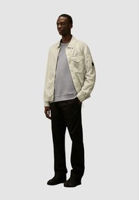 Lichtbeige gestreepte jas met voorzakken en een schouderpatch, gecombineerd met een grijze sweatshirts en zwarte broeken, gedragen met donkere schoenen.