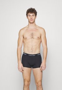 Boxer da uomo in cotone nero con una morbida vita che presenta il logo "Calvin Klein" in bianco. Il modello è senza maglietta, mostrando la vestibilità e il design.