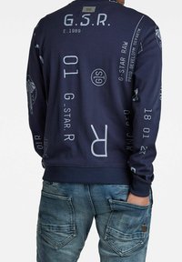 Sudadera de la marina con texto y logotipos en gris claro esparcidos. Puños y dobladillo acanalados. Combinada con jeans distendidos de color azul claro que presentan detalles en los bolsillos.