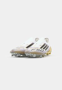 Bottes de football blanches avec des rayures noires, des accents dorés et un design texturé. Comprend des crampons pour une traction et une forme aérodynamique élégante.