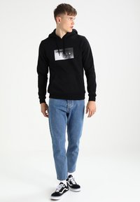 Zwarte hoody met een grafische print aan de voorkant, gecombineerd met lichtblauwe denim jeans en zwarte sneakers met witte accenten.