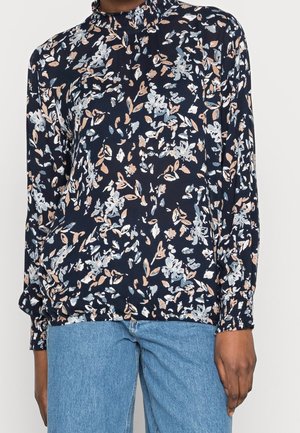 Blouse longue à manches longues à motifs floraux sombres avec un col montant à volants, portée avec un jean en denim bleu clair sur une personne visible du cou aux hanches.