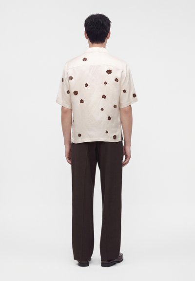 Homme portant une chemise beige à manches courtes avec un imprimé floral marron, un pantalon marron foncé et des chaussures foncées, debout de dos sur un fond blanc.