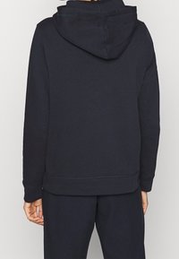 Granatowa bluza z kapturem o luźnym kroju, miękkiej fakturze i ściągaczach przy mankietach. Na plecach znajduje się minimalistyczny wzór bez widocznych logo.