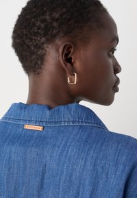 Denimhemd mit Kragen, versehen mit einem braunen Etikett auf der Rückseite; ein goldenes Creolen-Ohrloch betont das Ohr und hebt eine strukturierte Frisur hervor.