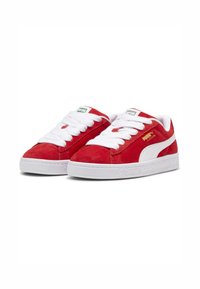 Röda mockasneakers med vita snören, med en vit sidstrimma och texturerad gummisula. Gyllene logotyp på utsidan.