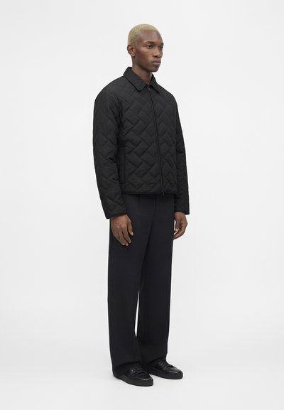 Άνδρας με κοντά ξανθά μαλλιά που φοράει μαύρο καπιτονέ μπουφάν με φερμουάρ, φαρδιά μαύρα παντελόνια και μαύρα αθλητικά, στεκόμενος πλάγια σε λευκό φόντο.