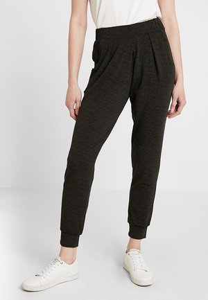Pantaloni jogger di un verde scuro, con texture, vita elastica, fronte plissettato e gambe affusolate. Abbinati a scarpe da ginnastica bianche.