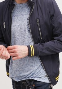 Giubbotto bomber della marina con polsini a strisce gialle, zip argentate e due tasche frontali, indossato sopra una T-shirt blu melange. Primo piano delle mani.