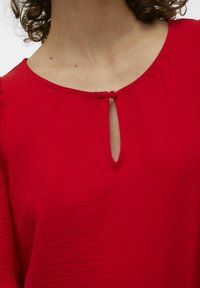 Rød bluse med rund halsudskæring og en nøglehulldetalje, fremstillet af blødt, tekstureret stof og med samlede ærmer.