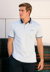 Světle modrá polo triko s námořnickým límcem a rukávovými lemy, se malým logem značky. Nosené s tmavými kalhotami, na pozadí pneumatik.