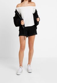 Haut blanc à épaules nues avec des détails en œillets, associé à un short noir avec des bordures en dentelle. Le modèle porte des baskets blanches et une veste noire.