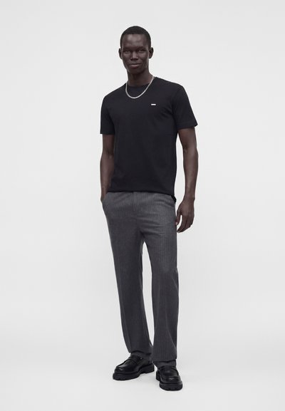 Homme debout les mains dans les poches, portant une chemise noire à manches courtes, un pantalon gris, des chaussures noires et un collier chaîne argenté.