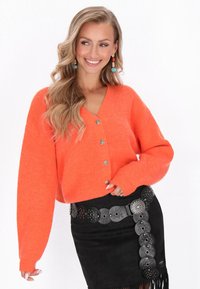 Pomarańczowy sweter o strukturze dzianinowej z dekoltem w kształcie litery V, z dużymi metalowymi guzikami, noszony na czarnej, zamszowej mini spódnicy z ozdobnym paskiem.