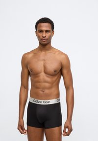 Crne bokserice s laganim sivim pojasom s logom Calvin Klein. Glatka tkanina, prilagođen dizajn, duljina do sredine bedra, minimalistički stil.