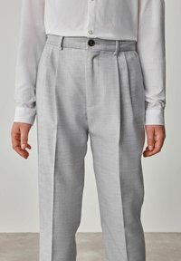 Pantaloni grigi chiari su misura con tessuto testurizzato, frontali plissettati e chiusura a un bottone, abbinati a una camicia bianca a maniche lunghe.