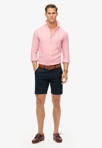 Camisa de lino rosa claro con botones en la parte delantera, pantalones cortos azul marino, cinturón marrón y zapatos de cuero marrón con cordones. El modelo se encuentra frente a un fondo blanco.