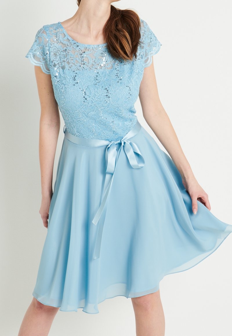 Femme portant une robe bleu clair avec un haut en dentelle à sequins, manches courtes, et un ruban en satin noué à la taille, tenant l'ampleur de la jupe.