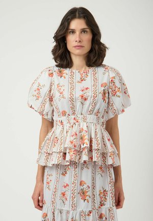 Mujer con un vestido blanco de flores con mangas abullonadas y detalles de volantes en capas, de pie frente a un fondo liso y claro.