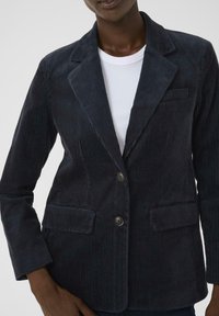 Marineblaue Corduroy-Blazer mit strukturierter Oberflächen, Ein-Knopf-Verschluss, Reverskragen und zwei unteren Taschen. Über einem weißen T-Shirt getragen.
