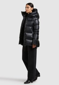 Chaqueta acolchada negra con un diseño brillante y acolchado y una capucha grande. Cuenta con una cremallera frontal y detalle de logo en la manga. Combinada con pantalones negros.