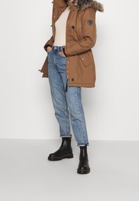 Brown parka jacket with fur-trimmed hood, zip and button closure, and side pockets. Worn with light blue jeans and black chelsea boots.