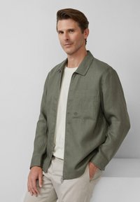 Lichte olijfgroene jas met een ritssluiting aan de voorkant, een borstzak en een relaxte pasvorm. Gepaarde met een beige shirt en lichte broek.