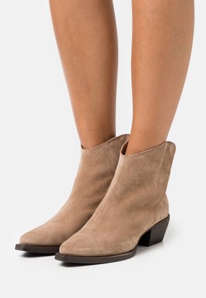 Jambes portant des bottines en daim brun clair avec des talons bas empilés sur un fond blanc.