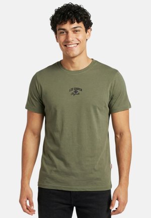 Jeune homme souriant aux cheveux courts et bouclés portant un t-shirt vert olive Lee Cooper Originals et un pantalon noir, debout devant un fond clair uni.
