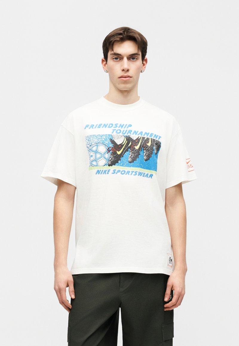 Junger Mann trägt ein weißes Nike Sportswear T-Shirt mit einem Fußballmotiv und dem Text "Friendship Tournament", kombiniert mit dunkler Hose.