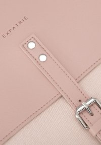 Correa de piel sintética rosa con herrajes plateados, unida a un fondo de lienzo texturizado beige, presenta la marca "EXPATRIÉ" en relieve.