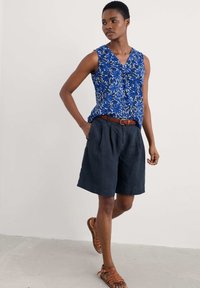 Blå blommönstrad ärmlös topp med V-hals, matchad med mörka lin shorts och bruna remsandaler. Texturerat bälte i accent.