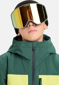 Gogle narciarskie z dużą, lustrzaną złotą soczewką, zieloną gumową oprawką i dopasowaną czarną czapką zimową. Kurtka jest zielona z żółtym poziomym paskiem.