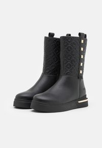 Bottes noires à la cheville avec une tige texturée présentant un motif de logo répétitif, une semelle plate et des carrés d'accent dorés sur le côté.