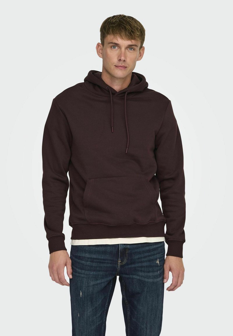 Sudadera con capucha color marrón con un bolsillo frontal, puños acanalados y material texturizado. Llevada sobre una camisa blanca y combinada con jeans oscuros.