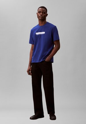 Jaunas vyras stovintis su viena ranka kišenėje, dėvintis mėlyną Calvin Klein marškinėlį, juodas kelnes ir juodus sportinius batelius prieš paprastą foną.