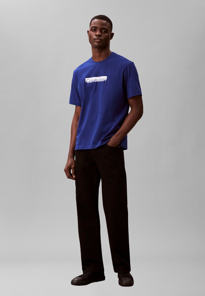 Νέος άνδρας που στέκεται με το ένα χέρι στην τσέπη, φορόντας μπλε μπλουζάκι Calvin Klein, μαύρο παντελόνι και μαύρα αθλητικά παπούτσια, σε απλό φόντο.