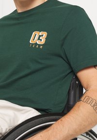 Zelené bavlněné tričko s bílou a zlatou grafikou a textem "03 TEAM". Krátké rukávy, regular fit a měkká textura.