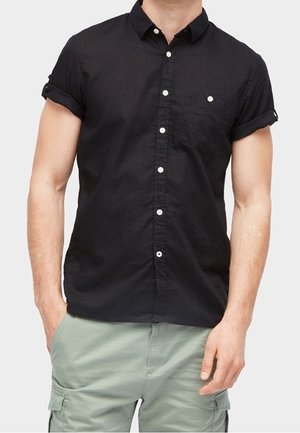 Homme portant une chemise noire à manches courtes avec poche poitrine et un short cargo vert clair, mains dans les poches, sur fond uni.