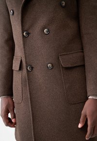 Brown wool double-breasted coat features black buttons, two front pockets, and a textured finish, shown at waist level with hands resting.