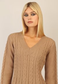Sweter z dzianiny w kolorze beżowym z dekoltem w kształcie litery V, z fakturowanymi warkoczami i długimi rękawami, wykonany z miękkiego materiału.