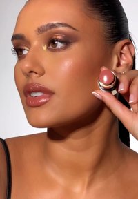 Runder, pfirsichfarbener Creme-Rouge-Compact mit glänzendem Deckel. Modell hält es nah an ihrer Wange und zeigt bronzefarbenes Make-up und glatte Haut.