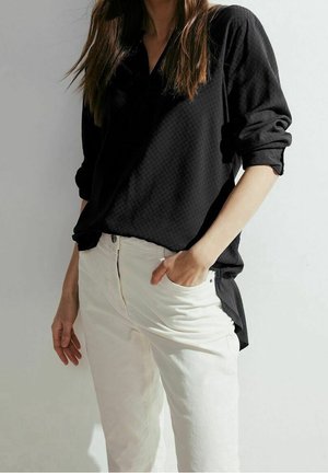 Vrouw draagt een zwart shirt met lange mouwen met opgerolde mouwen en witte broek, staat met één hand in de zak tegen een effen achtergrond.